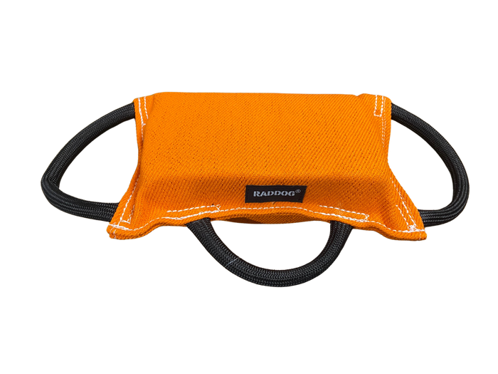 Bright orange padded dog training bite tug with a textured fabric surface, white stitching along the edges, a black “RADDOG” label at the center, and three black rope handles (two on the sides and one underneath), displayed against a black background.