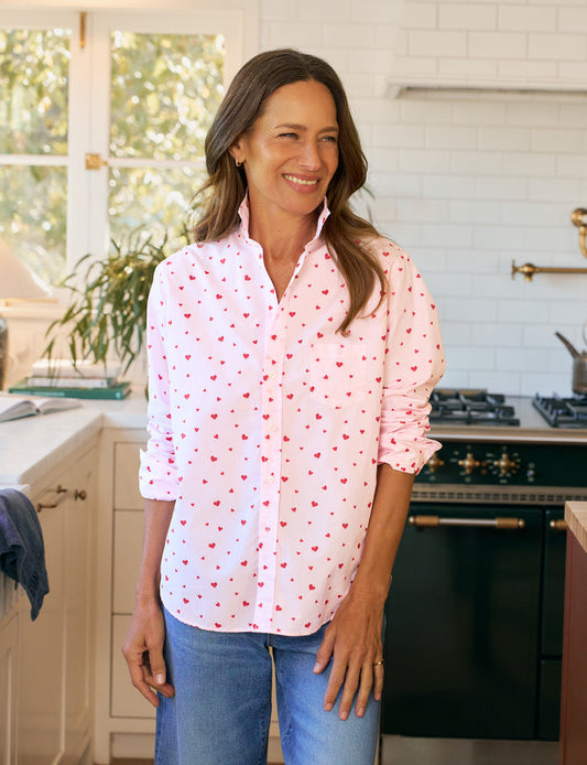 Woman standing in a kitchen wearing a pink shirt with red hearts and blue jeans.