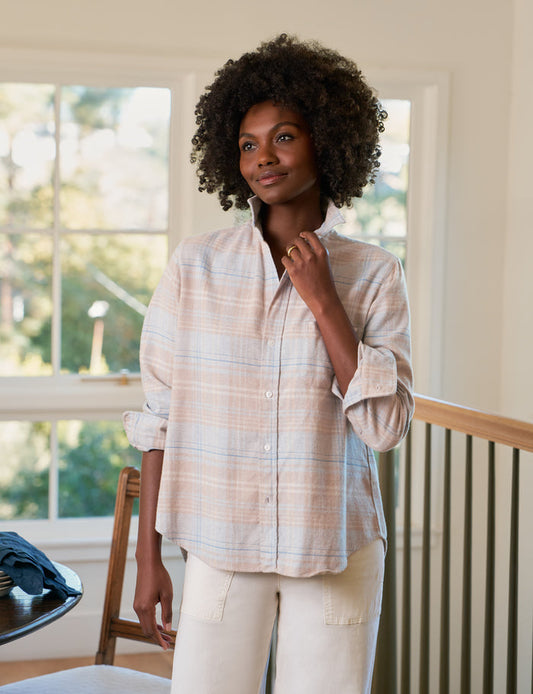 Woman wearing a plaid shirt and white pants standing in a room with large windows.