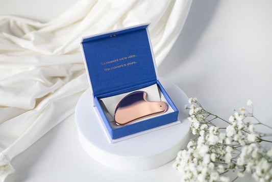 A luxurious golden acupressure facial lift tool by LILIXIR, displayed in an open blue box with a reflective surface, accompanied by a white floral arrangement and soft fabrics in the background.