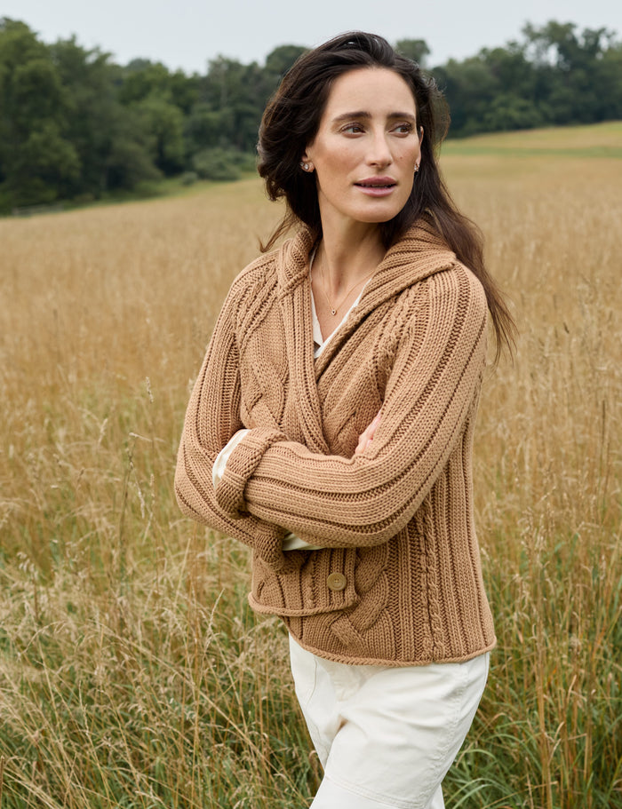 A woman stands in a field of dry golden grass wearing a chunky cable-knit brown hooded cardigan and white trousers.