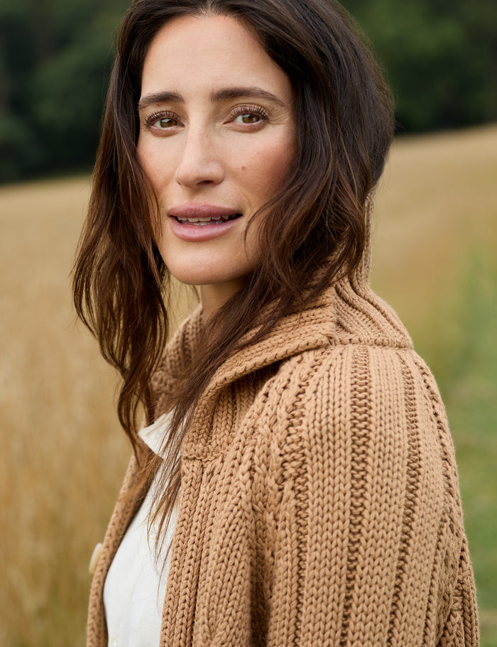A woman with long brown hair wearing a chunky knit beige cardigan over a white top, photographed outdoors against a blurred field background.