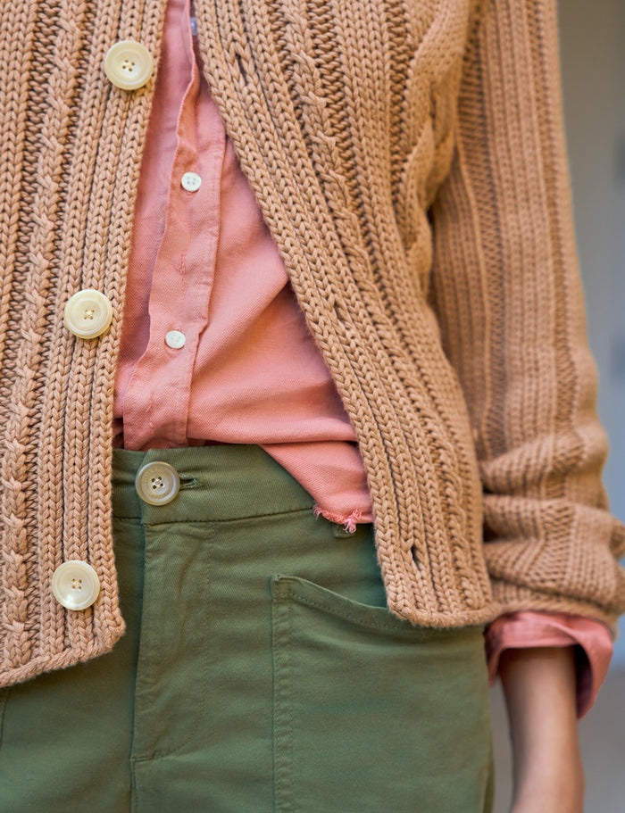 A close-up of a person wearing a chunky knit cardigan in a warm tan/beige color over a pink button-up shirt and olive green pants.