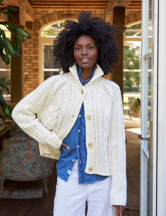 A woman standing in a doorway wearing a beige, cropped cable knit cardigan with horn buttons over a blue shirt and white pants.