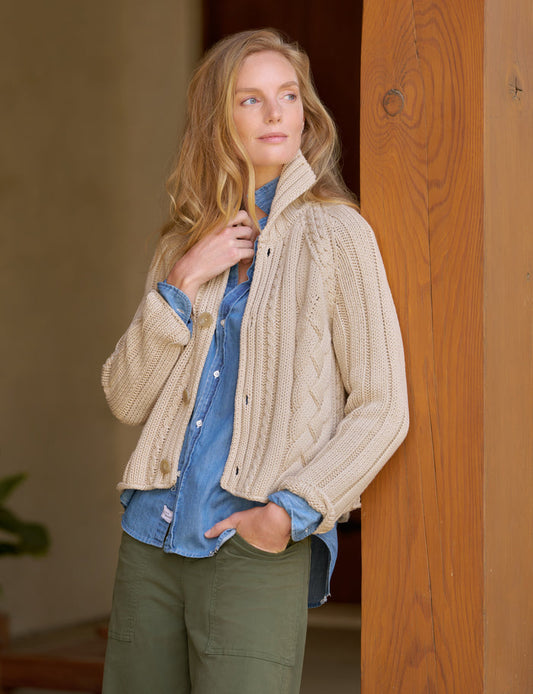 A woman wearing a cream cable-knit button-front cardigan over a blue denim shirt and olive green pants, standing next to a wooden door frame.