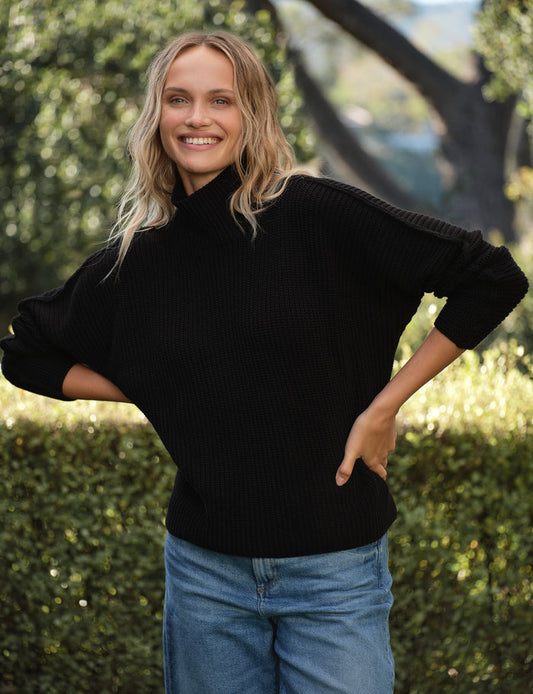 Woman wearing a black sweater and blue jeans standing outdoors with greenery in the background