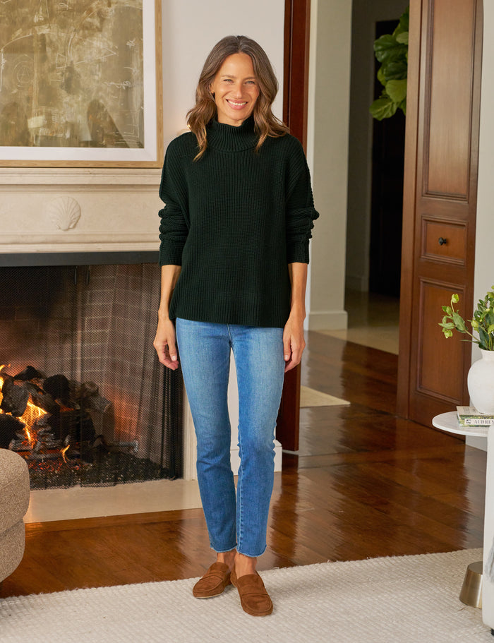 Woman wearing a dark green sweater and blue jeans standing in a living room with a fireplace.