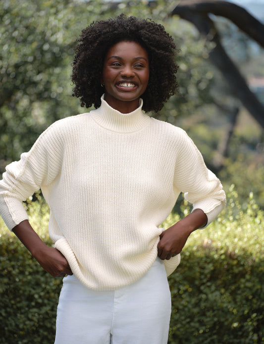 Woman wearing a white sweater and white pants standing outdoors with greenery in the background