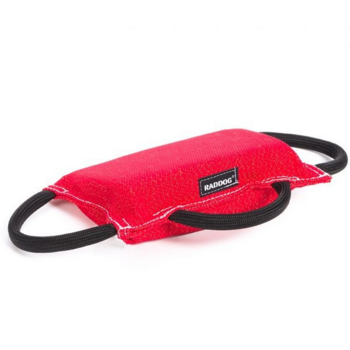 Red padded dog training bite tug with a textured fabric surface, white stitching along the edges, a black “RADDOG” label on the side, and two black rope handles on each end, displayed against a light background.