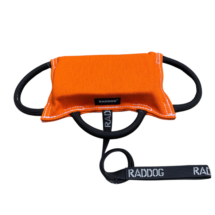 Orange dog training bite pad made from durable fabric, featuring reinforced stitching, multiple black loop handles, and a RADDOG logo label, displayed on a white background.