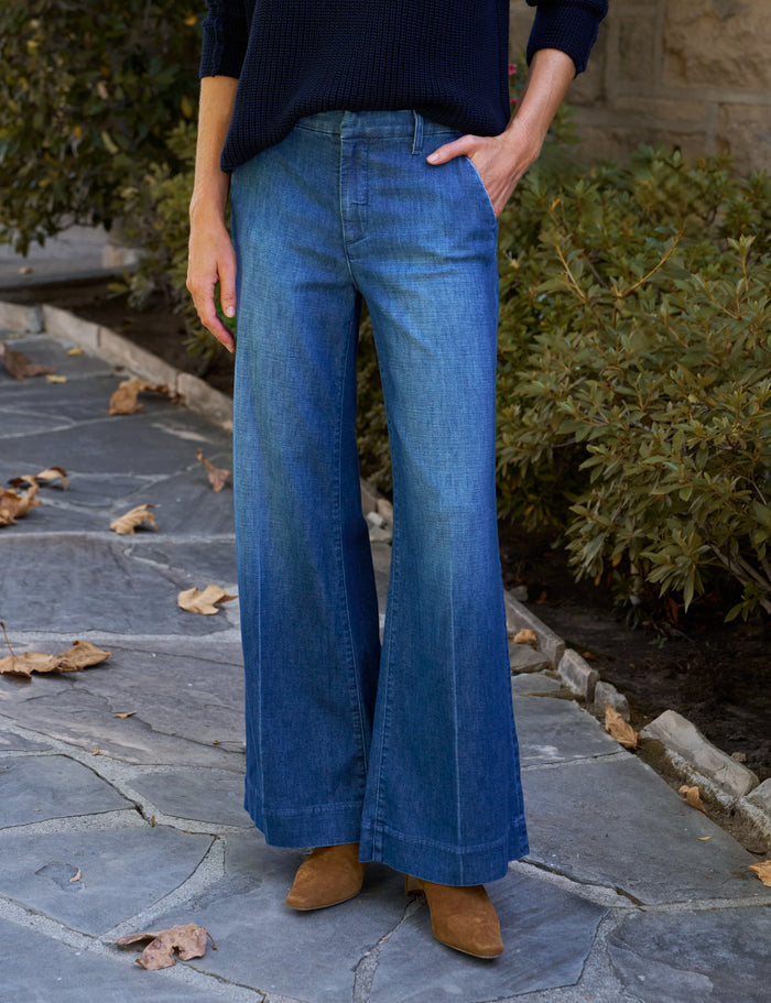 A person wearing blue wide-leg denim jeans, a dark navy ribbed sweater, and tan ankle boots, standing on a stone patio with green shrubs in the background.